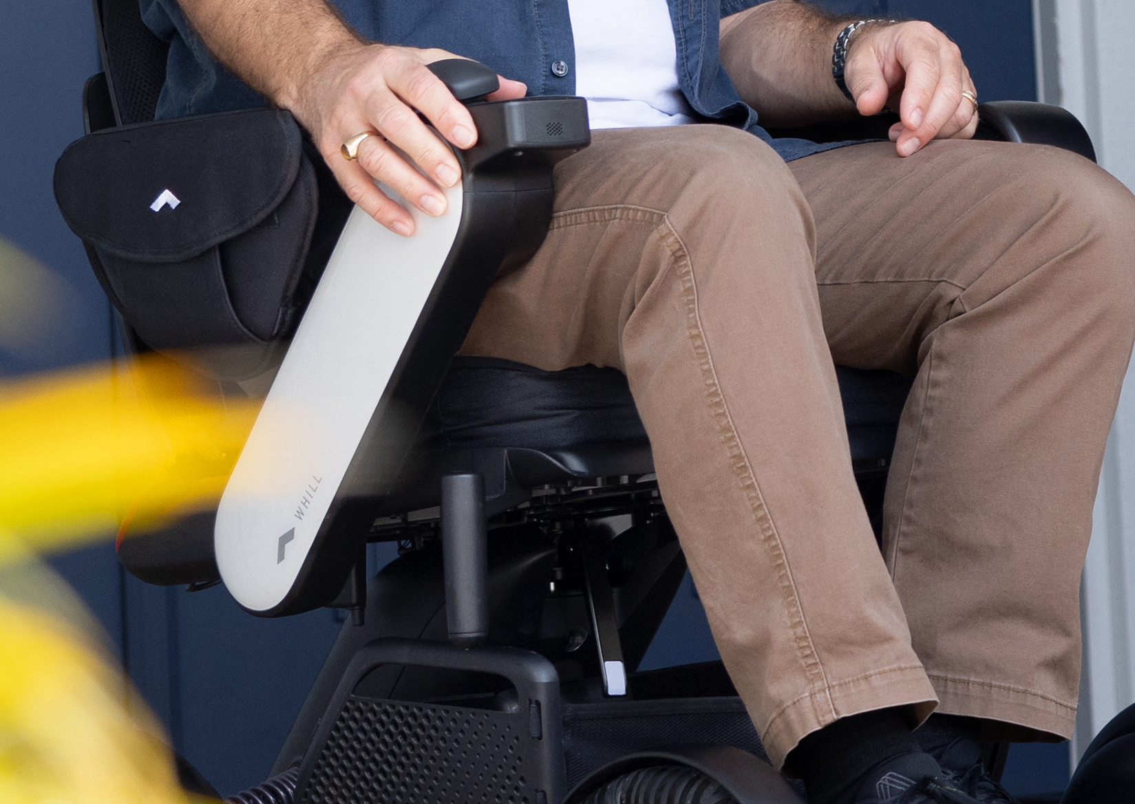 A man seated in a WHILL Model C2 Power Chair, wearing beige pants and a blue shirt, with one hand resting on the armrest controller.