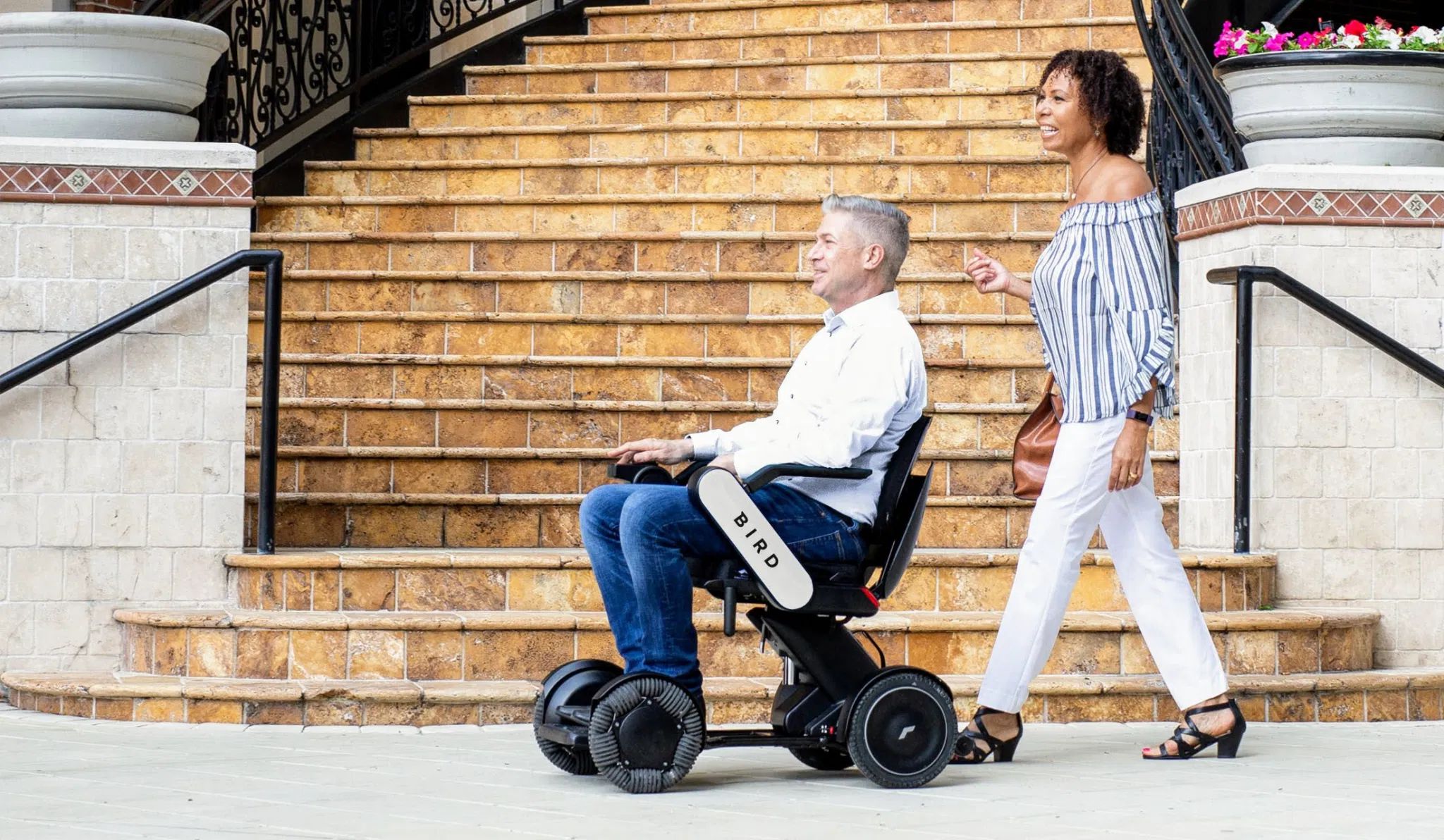 A man in a WHILL Model C2 power chair smiles while riding beside a woman walking. They are on a sidewalk in front of wide stone steps, creating a positive and inclusive atmosphere.