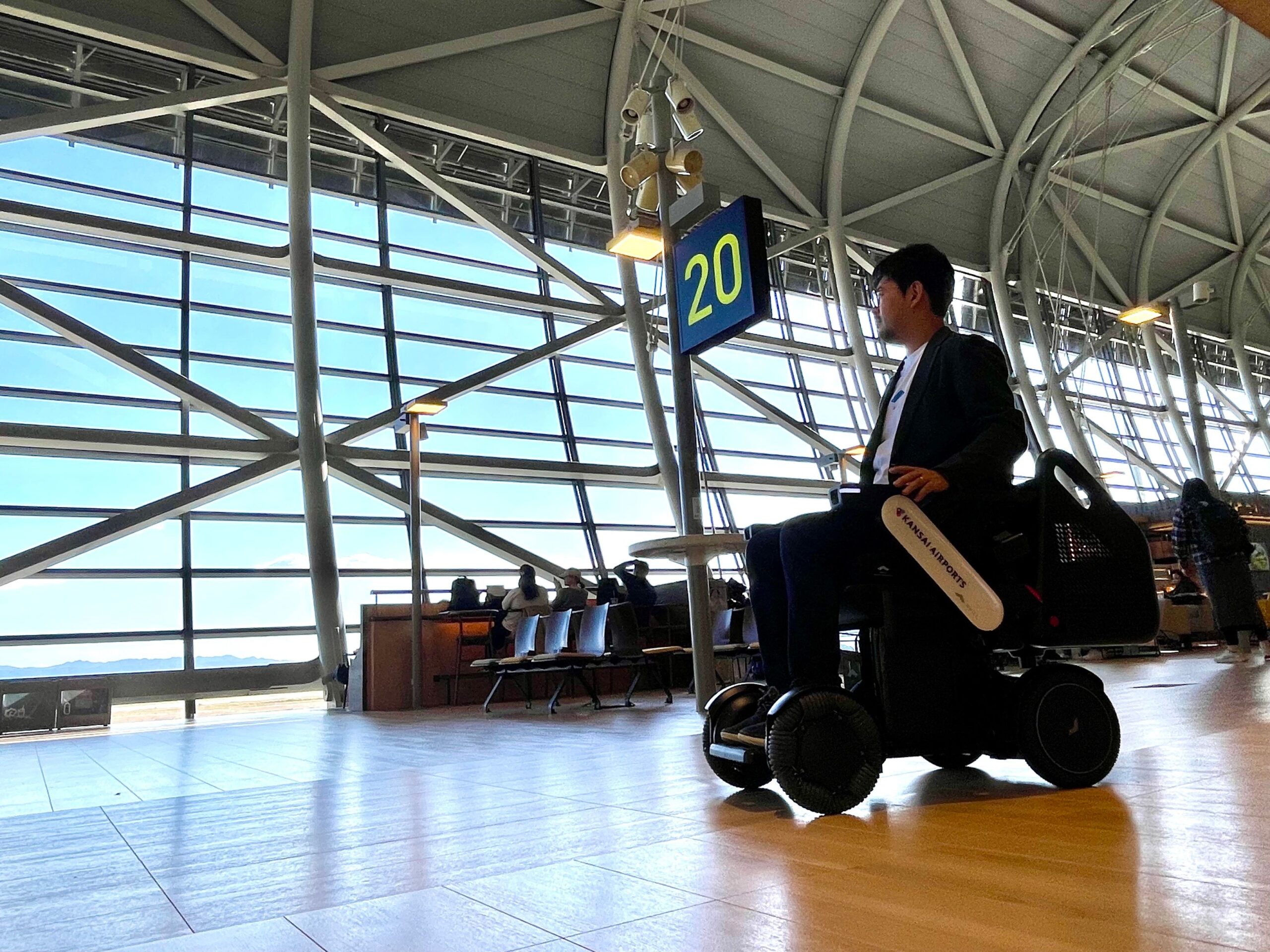 Man in a WHILL autonomous service power chair in an airport terminal, gazing outside through large windows.