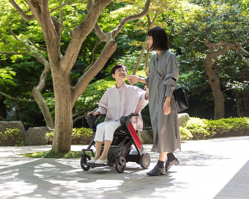 Two women enjoying a sunny day in a park. One woman is in a WHILL Model F folding power chair smiling, while the other stands beside her, both wearing stylish dresses.