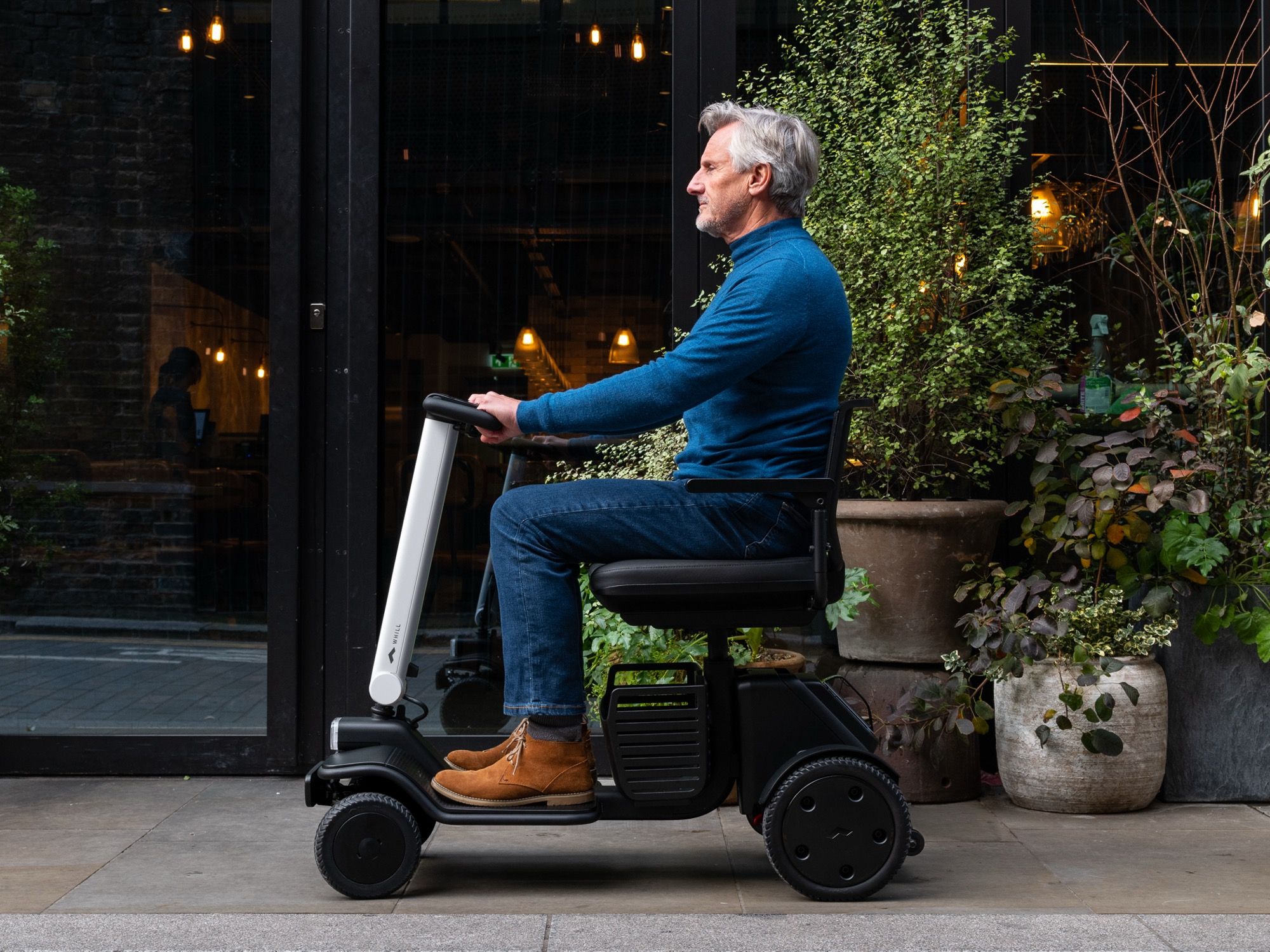 A man in a blue sweater and jeans sits on a WHILL Model R mobility scooter. He is side-on, beside large potted plants, conveying a relaxed and calm atmosphere.