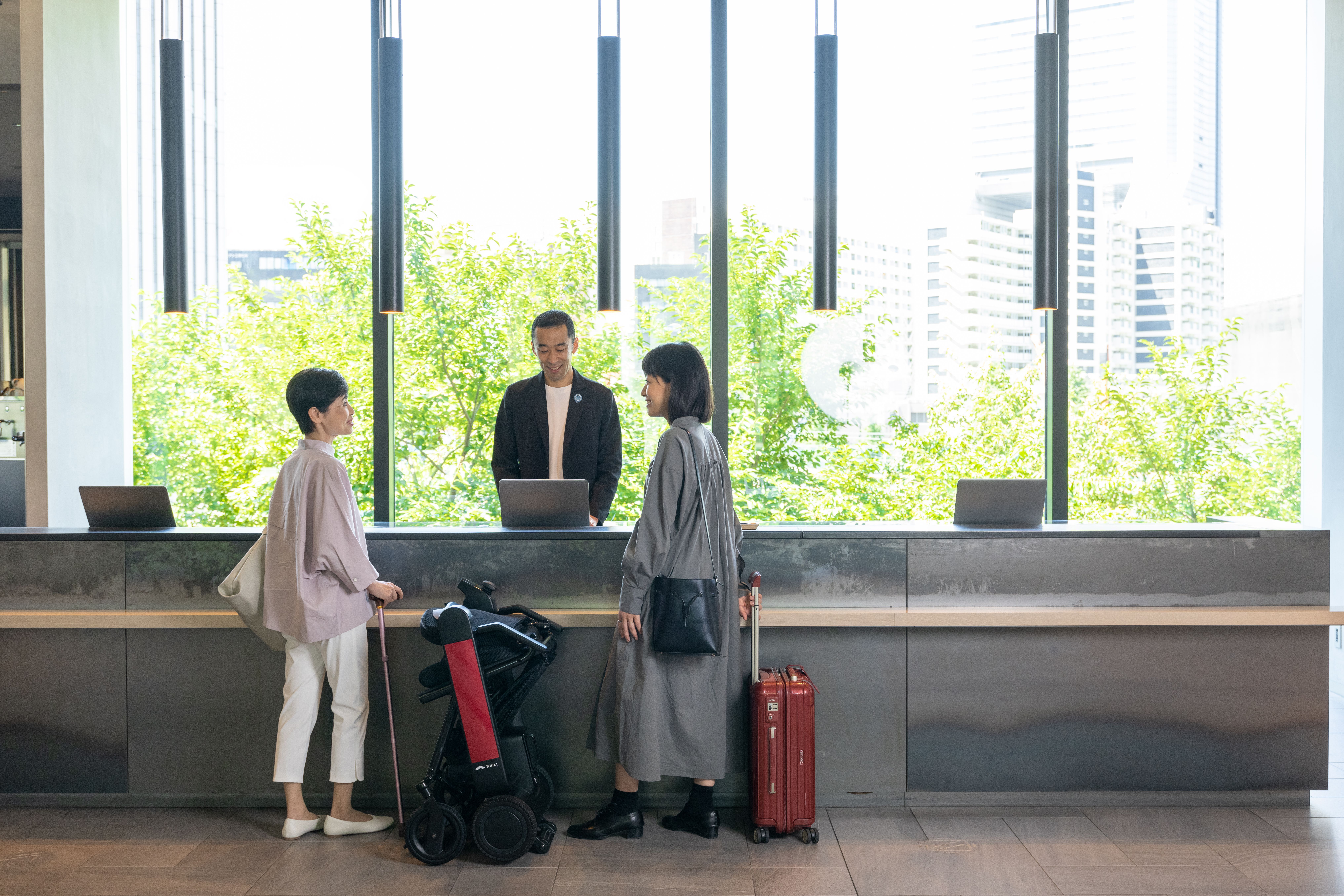 Two people with luggage and a Model F power chair engage with a receptionist at a modern hotel lobby, with large windows and city views. The atmosphere is professional and welcoming.