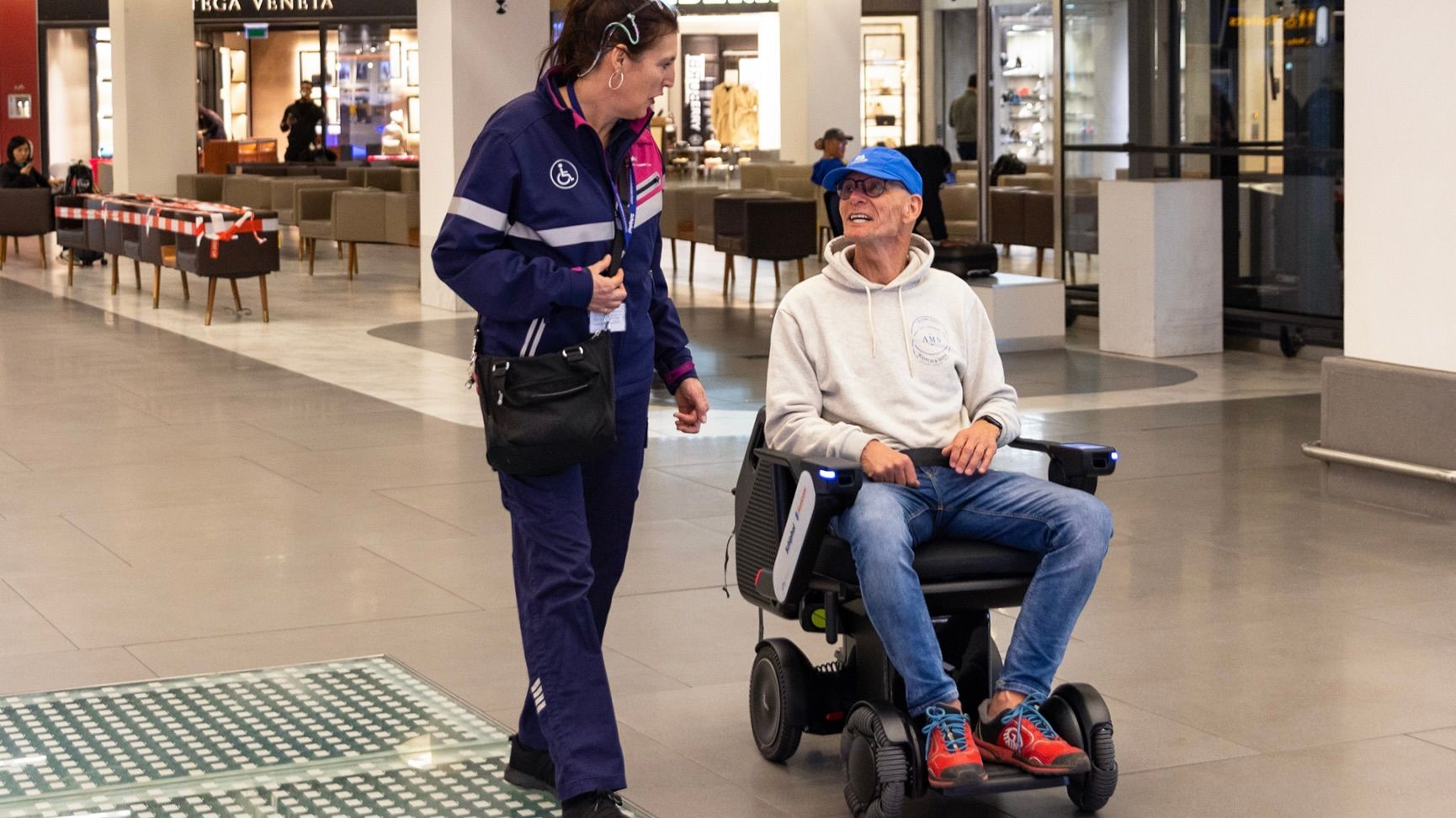 A smiling man in a blue cap and hoodie sits in a WHILL autonomous service power chair, chatting with a woman in a purple uniform.