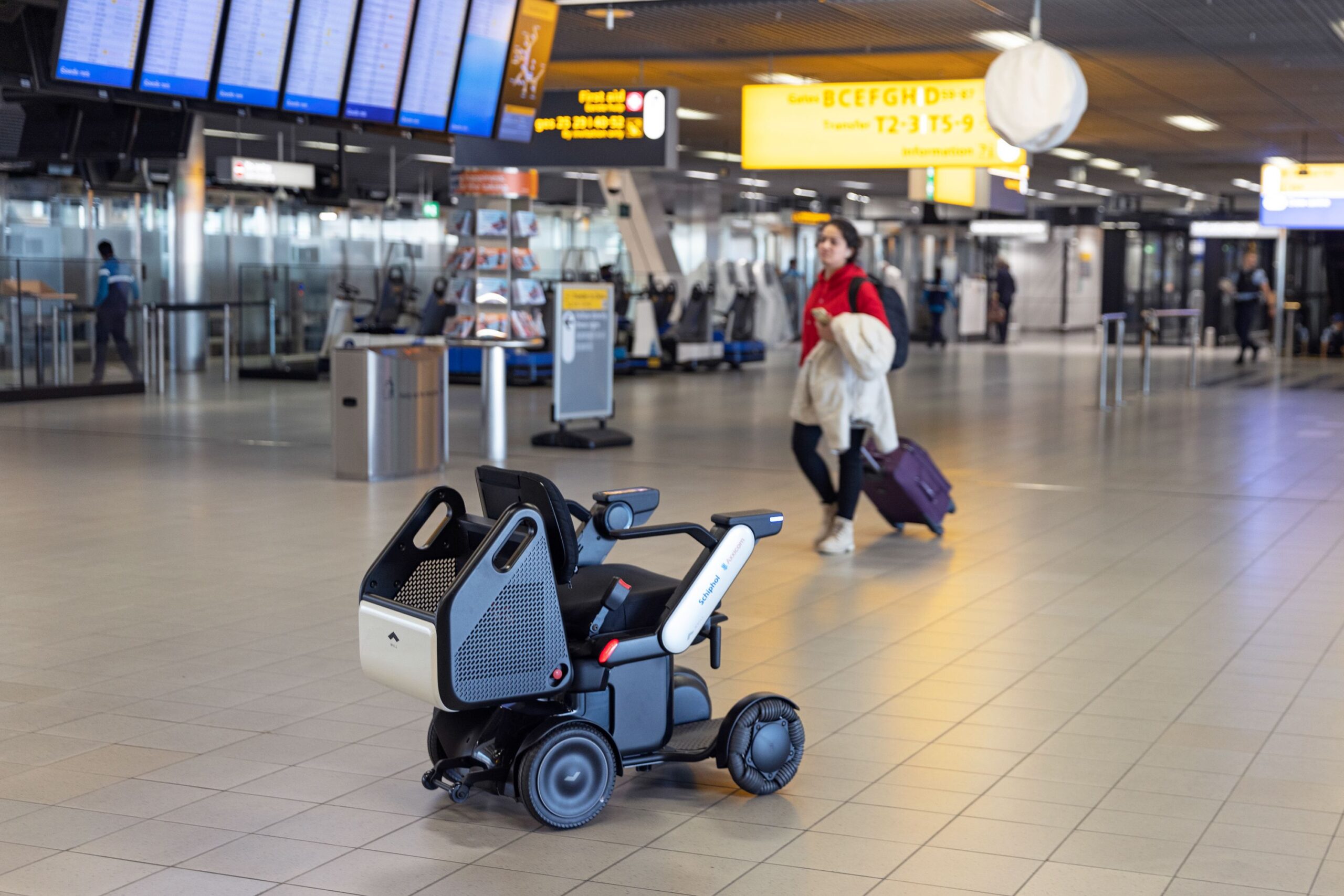 A WHILL Autonomous service power chair sits in the center of a spacious airport terminal. In the background, a traveler with luggage moves past.