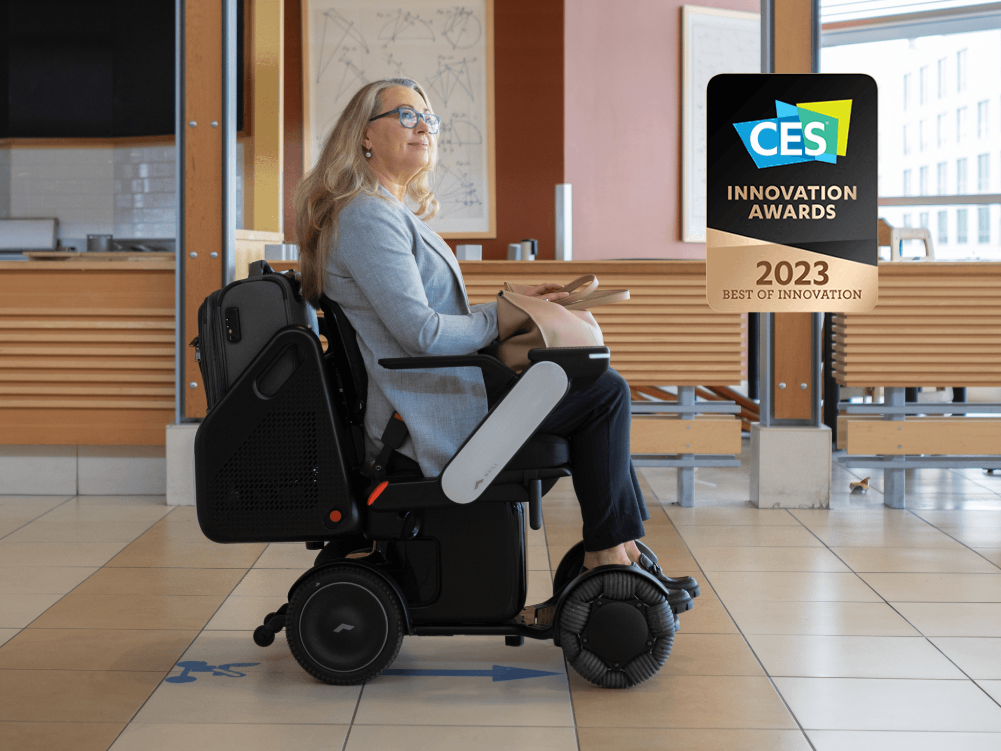 A woman in a WHILL autonomous service power chair, with a CES 2023 Innovation Award badge, smiles confidently indoors, surrounded by sleek, minimalist decor.