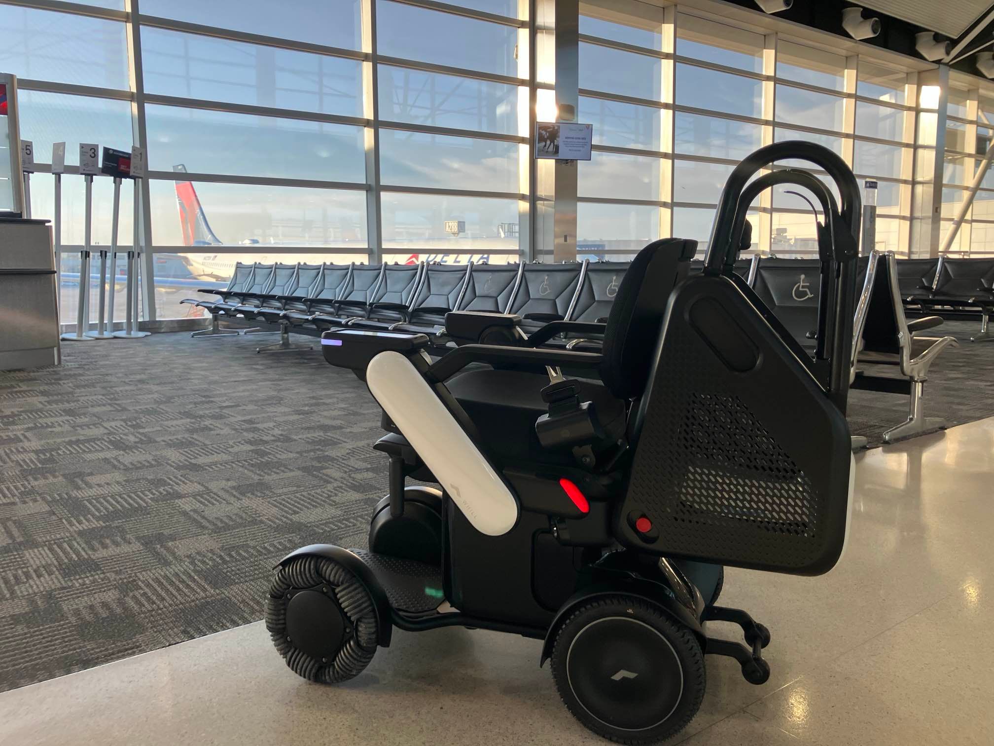 A WHILL Autonomous Service Power Chair is positioned in a sunlit airport terminal, near rows of empty seats and large windows overlooking airplane tails.
