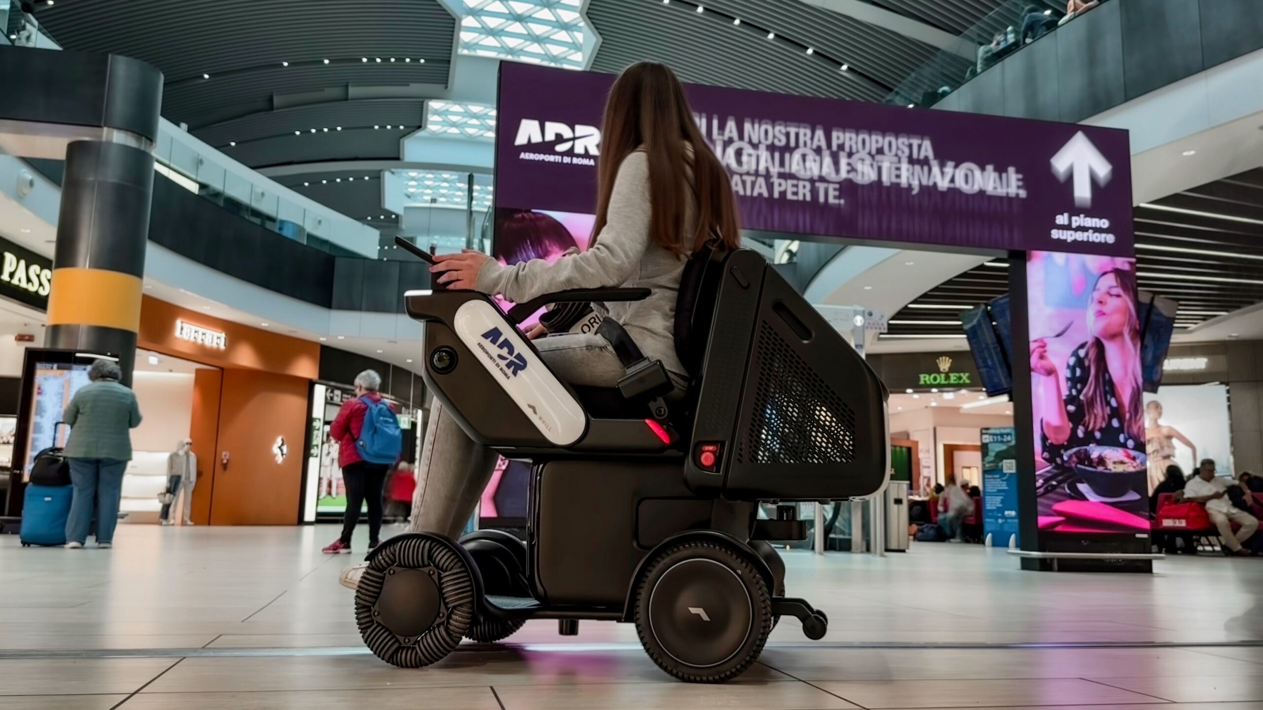 A person in a WHILL autonomous service mobility device navigates an airport, surrounded by shops and digital signage. The scene is busy, conveying travel and innovation.