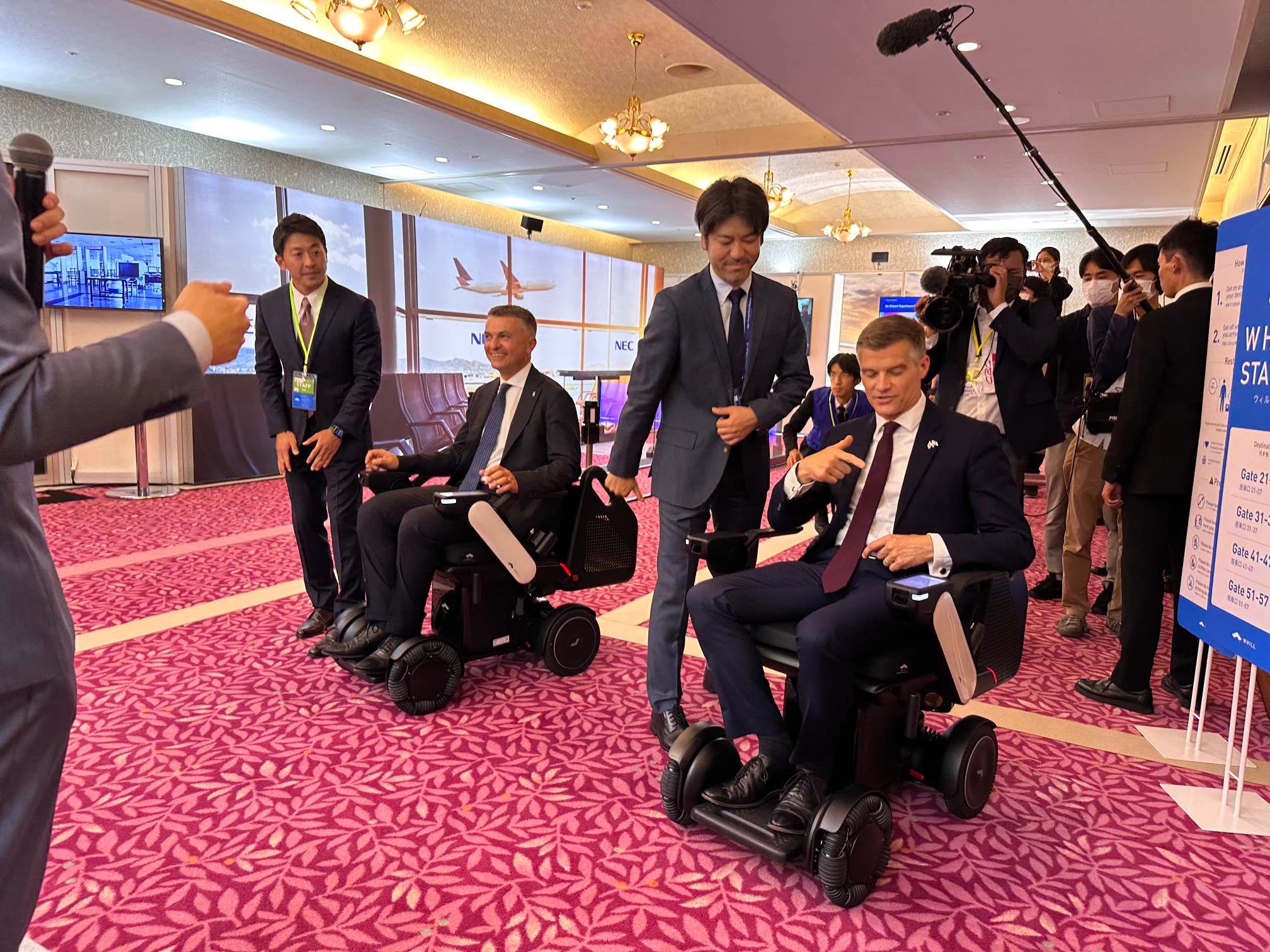 Men in suits sit in WHILL autonomous service power chairs on a pink carpet, surrounded by journalists in a conference setting. The mood is professional and engaged.