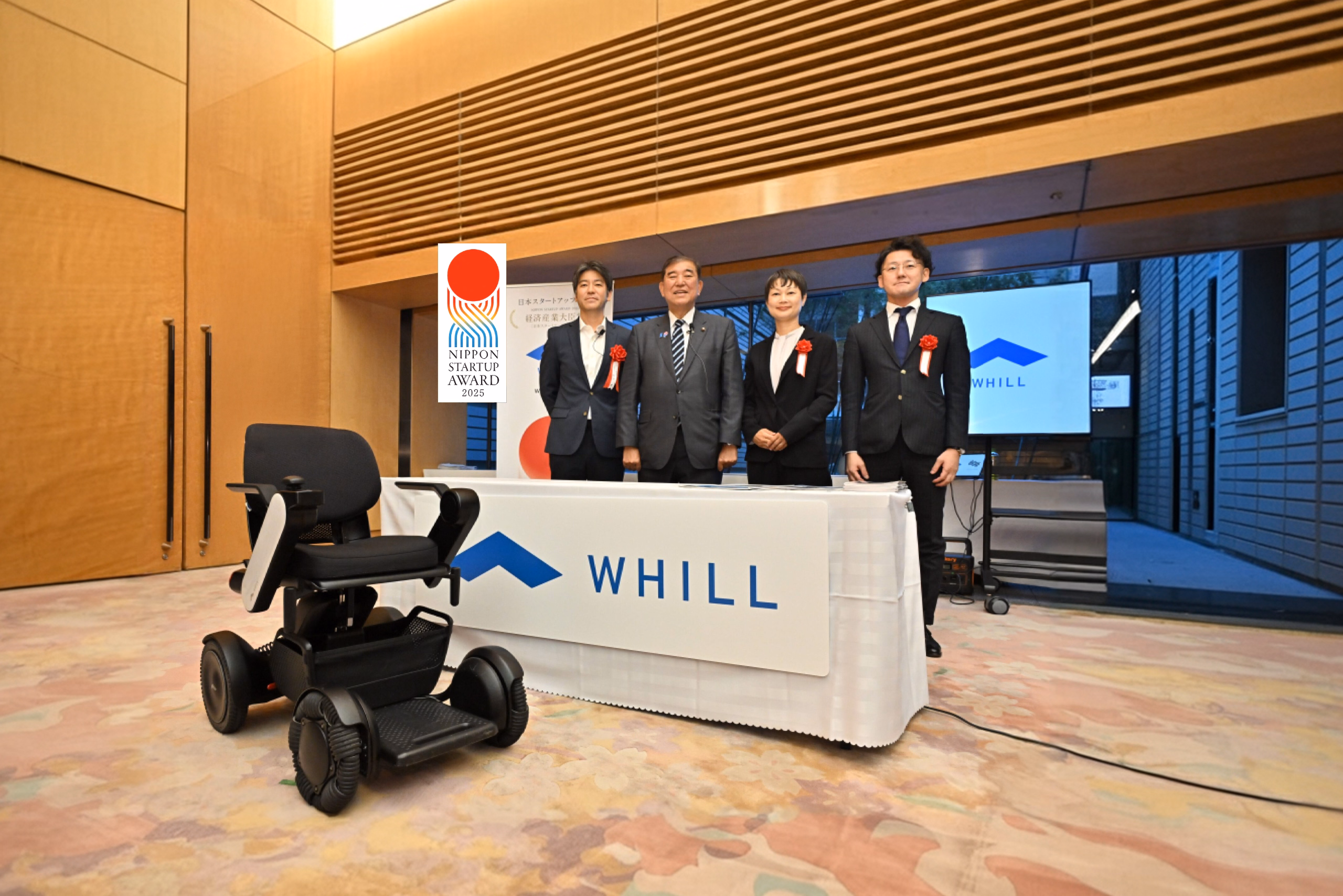Four people stand smiling behind a table with "WHILL" on it. A black mobility chair is in the foreground. A "Nippon Startup Award 2025" sign is visible.
