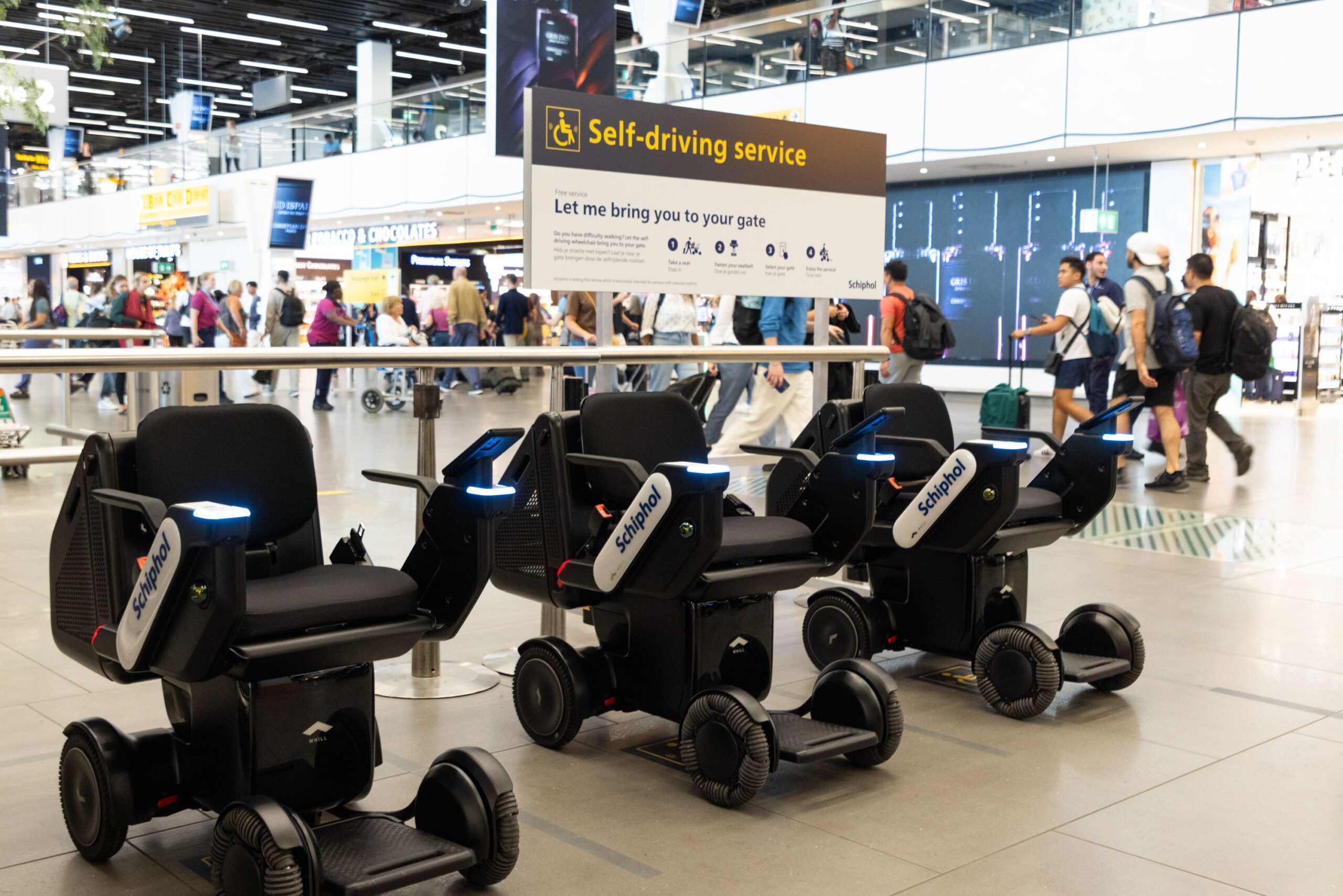 Three self-driving wheelchairs at an airport with a "Self-driving service" sign above. People walk by in the background, creating a busy atmosphere.