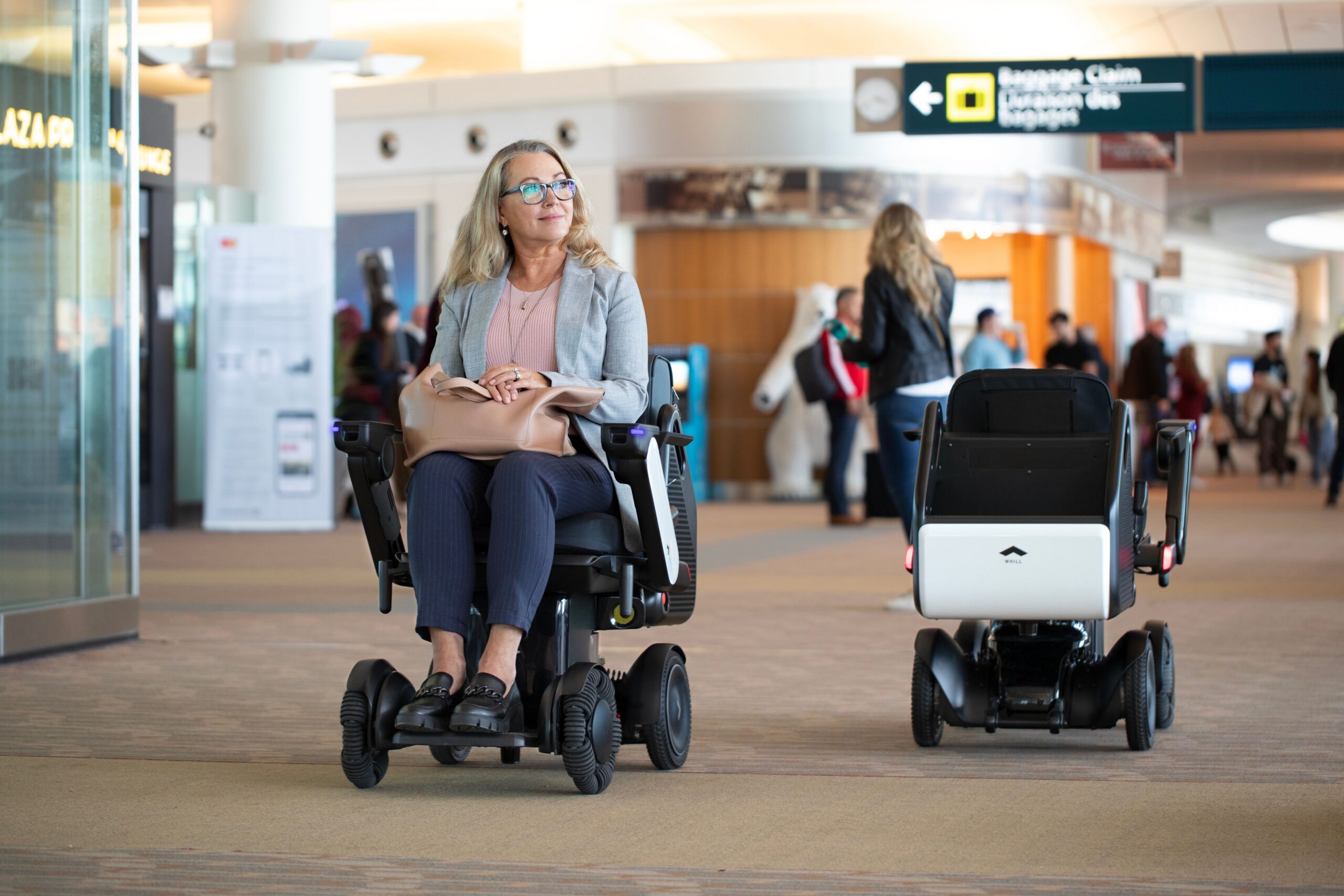 A woman seated in an WHILL Autonomous service power chair in an airport, looking content and holding a bag. Another unoccupied wheelchair is nearby. People and signs are in the background.