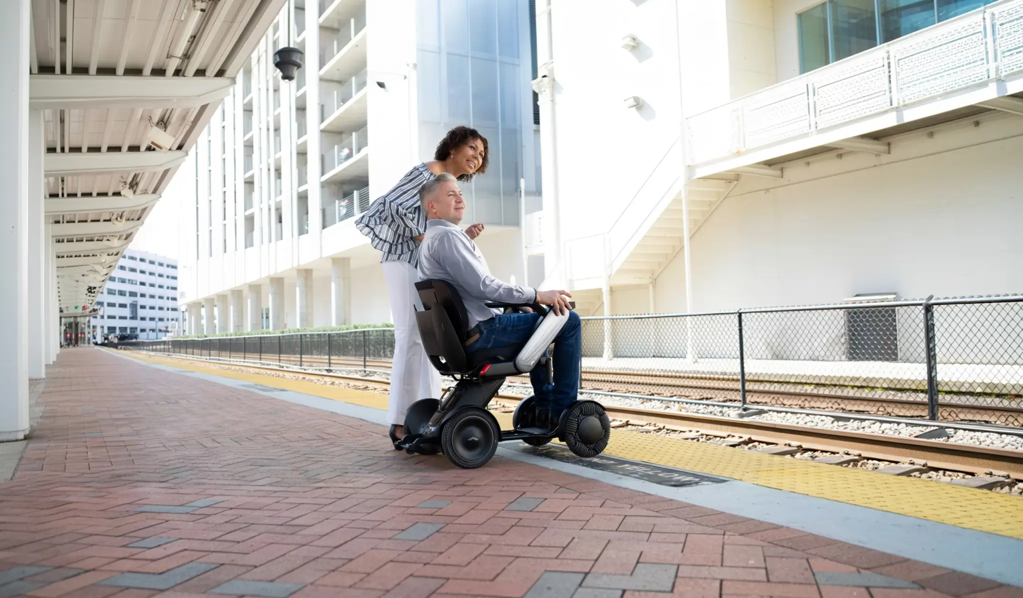 Women standing next to a man sitting in a WHILL Model C2 powerchair at a train station.